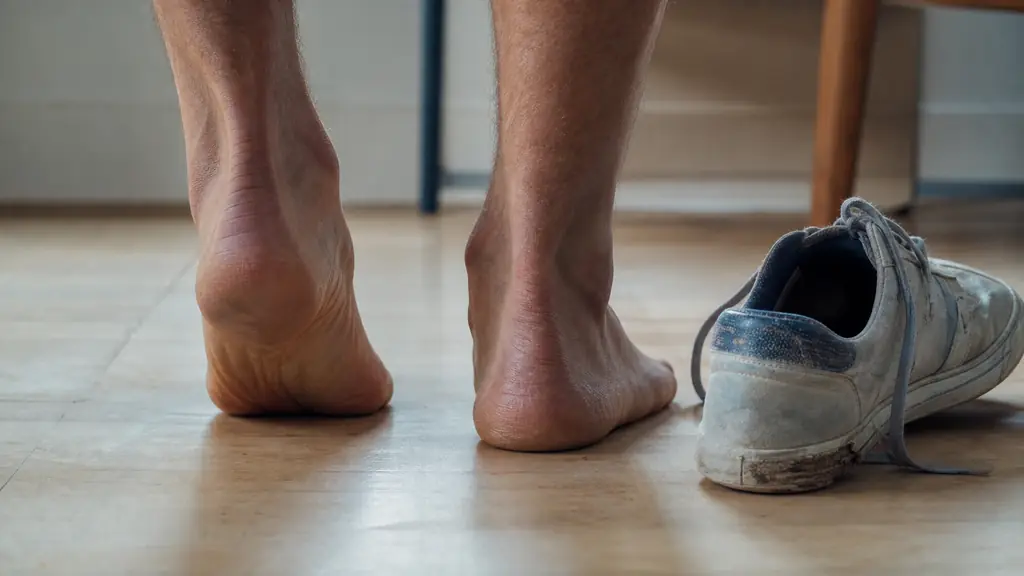Editorial photo of bare adult feet standing on a light floor, viewed from behind, showing one foot with a very visible inner arch and the other flatter, with a worn sneaker beside them showing outer-edge sole wear, no text, no logo