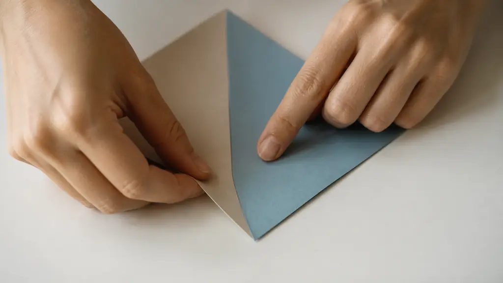 Close-up editorial photograph of hands aligning two corners of a square two-tone origami sheet on a flat table, one fingernail carefully sharpening the central fold