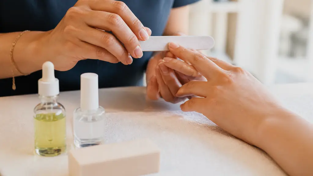 Editorial close-up of an esthetician filing a natural fingernail beside a cuticle oil bottle, base coat and nail buffer on a clean manicure table, no text, no logo