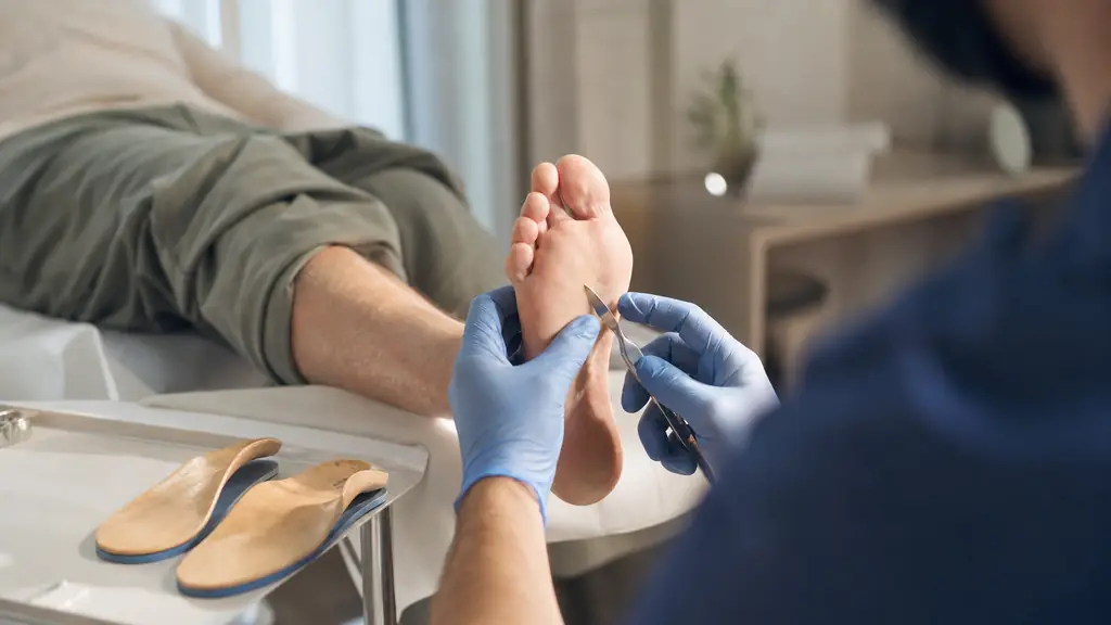 Close-up editorial photograph of a podiatrist examining a patient's foot on a treatment table, with a podiatric instrument for callus care and a pair of custom plantar orthoses visible nearby, no text, no logo