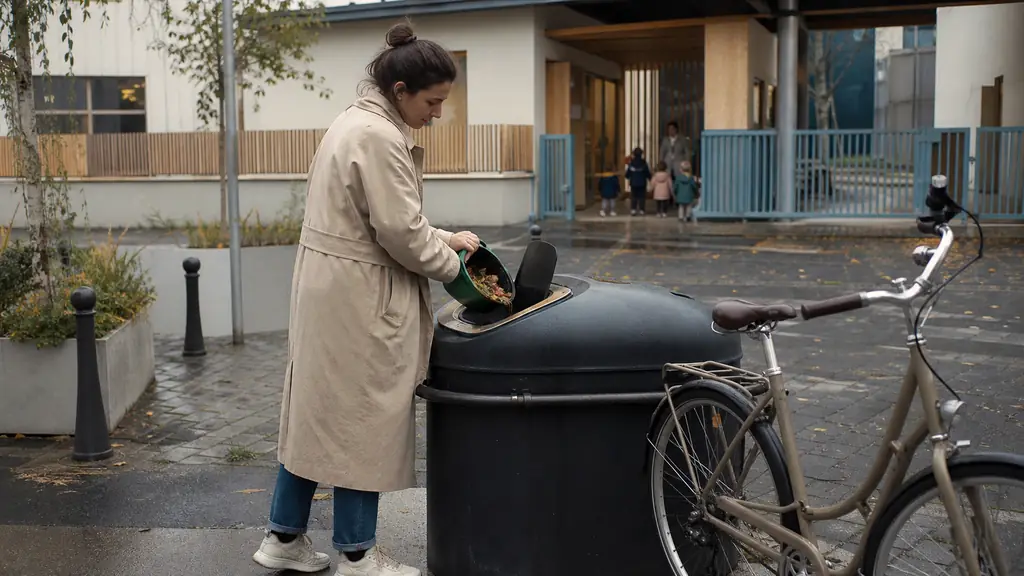 editorial photograph of a resident placing food scraps into a communal biod waste collection point beside a bicycle, with a childcare center entrance in the background, no visible text or logos