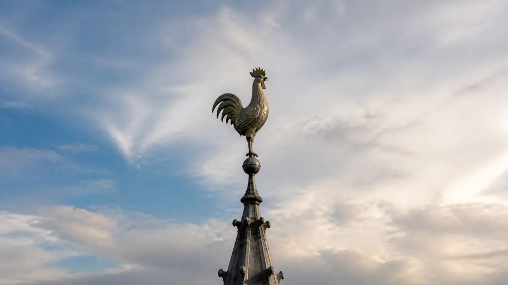 Editorial photograph of the rooster weather vane crowning the spire of Notre-Dame Cathedral, silhouetted against the Paris sky, with its metal body and directional position clearly visible, no text, no logo