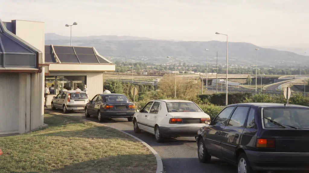 1990s roadside fast-food restaurant on the outskirts of a French town, cars entering a drive-through lane, motorway interchange and Rhône valley landscape in the background, no text, no logo