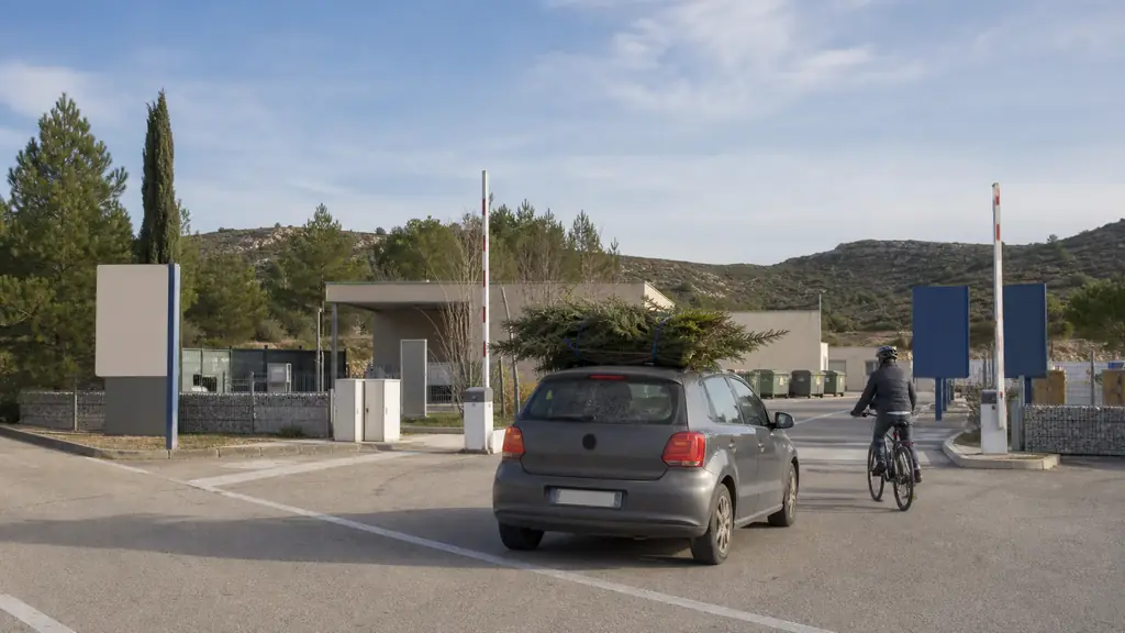 Editorial photograph of a waste collection facility entrance reached from the D611 in southern France, with a car carrying garden waste and a cyclist approaching, Mediterranean Corbières landscape, no visible text or logo