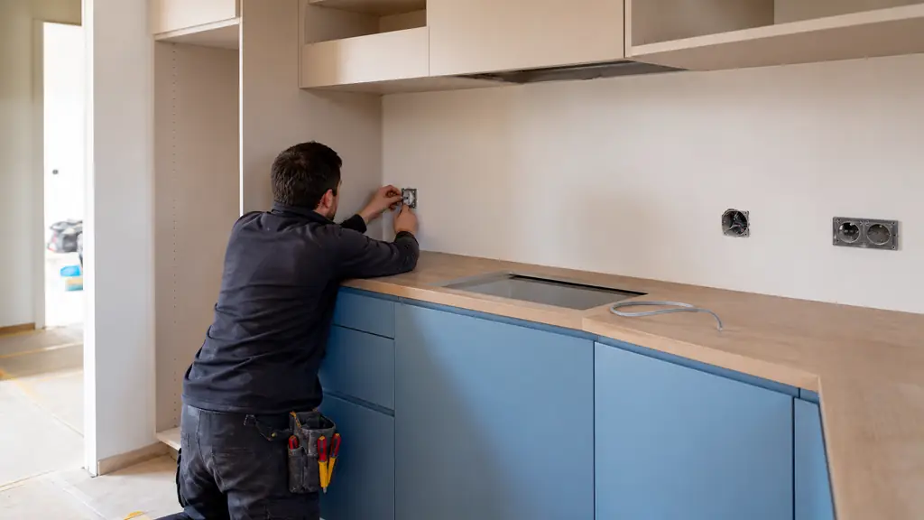 Editorial photograph of kitchen renovation in progress near Paris, showing newly painted cabinet fronts, a fitted laminate worktop and an electrician installing a wall outlet, natural daylight, no text, no logo