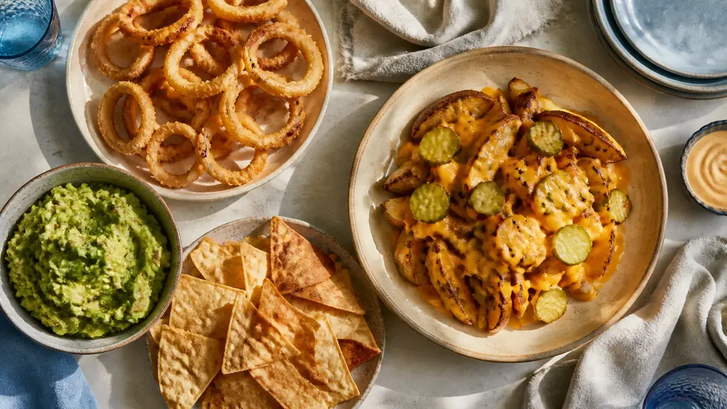 Overhead editorial photograph of a casual American snack table with crispy onion rings, cheddar-covered potato wedges with pickles, and a bowl of chunky guacamole beside nachos, natural evening light, no text, no logo