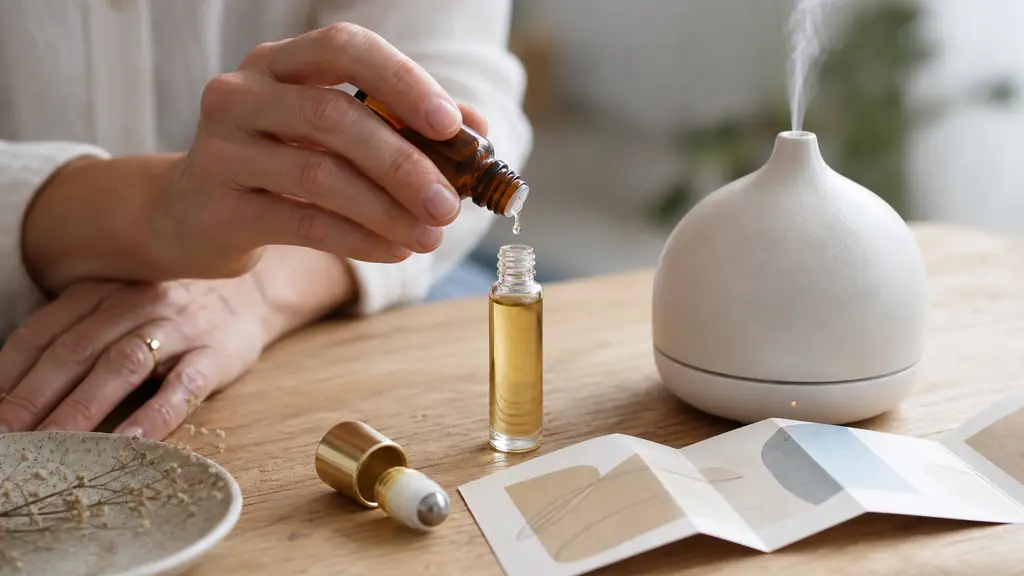 Editorial photograph of a hand adding one drop of neroli essential oil to a 10 ml jojoba roll-on beside a small ultrasonic diffuser and an open product label on a light wooden table