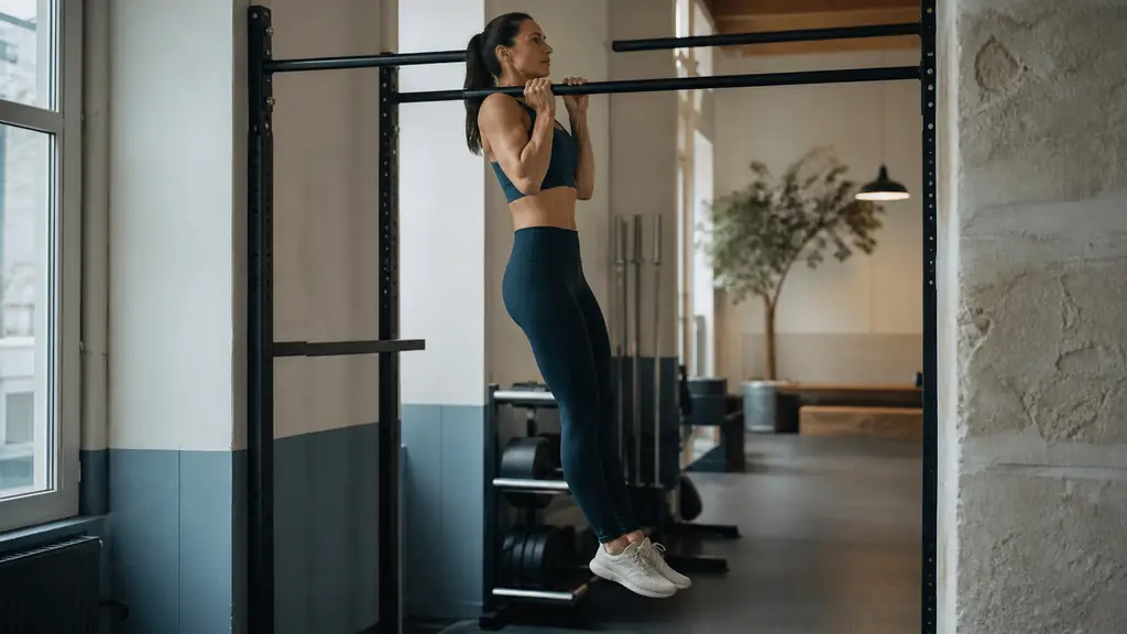 Editorial fitness photograph showing a side view of a woman practicing a controlled supinated chin-up, elbows close to the torso, wrists aligned and legs still, natural gym lighting, no text or logo