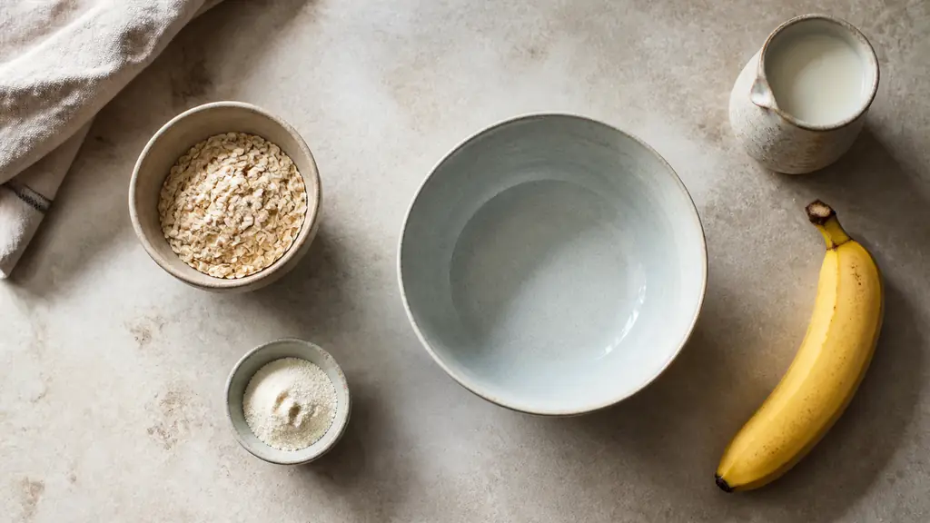 Overhead editorial photograph of rolled oats, half a scoop of whey protein, almond milk and a ripe banana arranged beside a mixing bowl on a natural kitchen worktop, soft morning light, no text, no logo