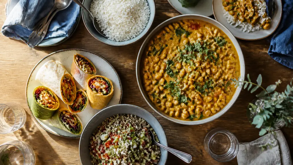 Overhead editorial photograph of chickpea coconut curry beside a bowl of rice, colorful hummus wraps and a quinoa salad with feta on a family kitchen table, natural daylight, no text, no logo