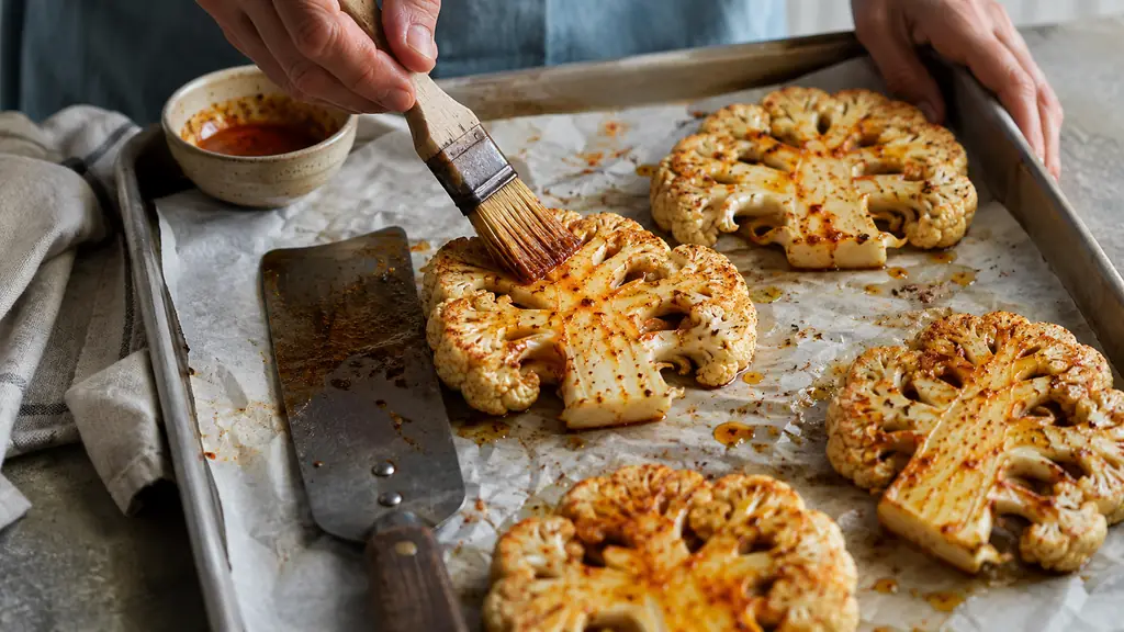 Hands brushing paprika oil onto thick central cauliflower slices with the core intact, arranged on a parchment-lined baking tray, wide spatula beside them, natural kitchen light, editorial food photography, no text, no logo