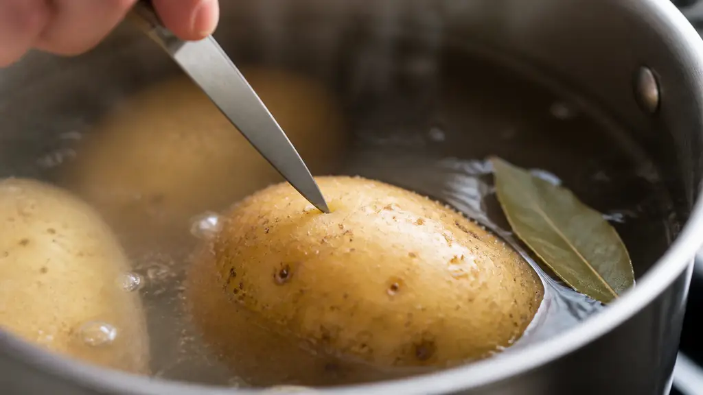 close-up editorial food photograph of a knife tip testing a skin-on potato in a gently simmering saucepan, with bay leaf and visible small bubbles, no text or logo