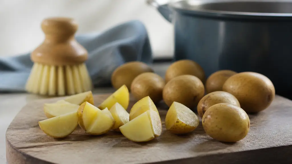 editorial food photograph of equal potato quarters beside whole skin-on baby potatoes on a wooden board, with a vegetable brush and saucepan in the background, no text or logo
