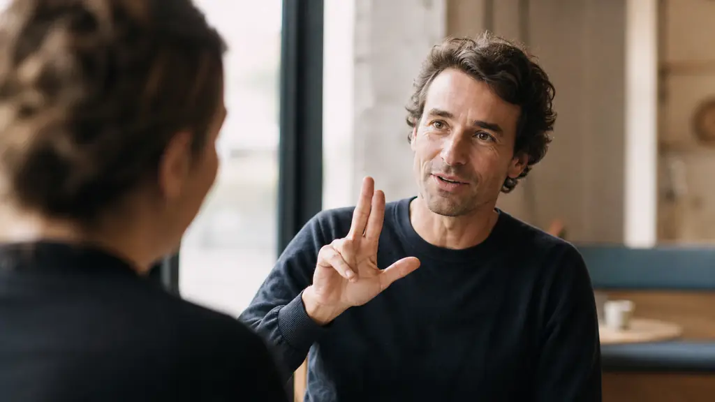 right-handed adult slowly fingerspelling a proper name at chest height while maintaining eye contact with a deaf conversation partner, relaxed arm, expressive face, natural editorial photography, no text, no logo