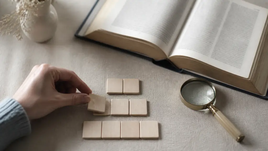 Editorial photograph of a hand arranging blank wooden letter tiles into rows of two, three and four beside an open dictionary and a small magnifying glass, no readable text, no logo